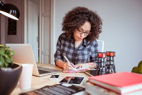 Female freelancer works from home office, writing in a notebook in front of a laptop