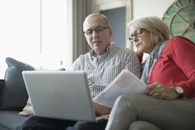 Senior couple with paperwork using laptop on living room sofa