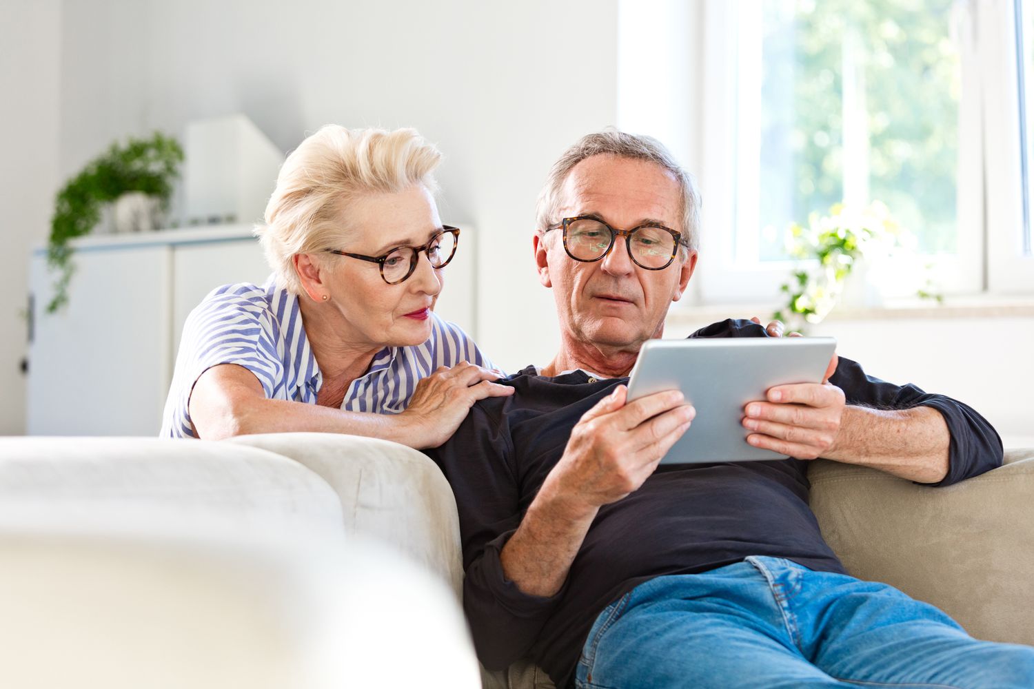 Elderly man sitting on sofa in the living room at home and showing something on digital tablet his wife.