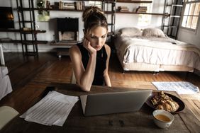 Woman working on laptop at home with pastries and coffee, chin in hand