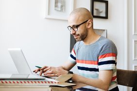 Bald man with glasses sits at desk with laptop and pen in hand