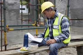 Builder eating packed lunch looks at documents on construction site