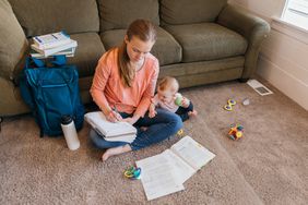 Mother working with a paper notebook on living floor, infant beside her