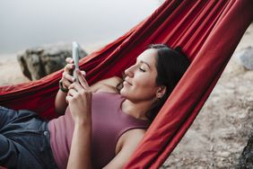 A woman laying in a hammock looks at a phone.