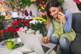 Female flower shop owner on cell phone looking at computer while sitting near flowers