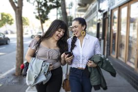 Two women walking down the street laughing while looking at a phone together