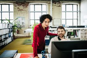 Woman standing behind coworker who is sitting at his desk. Both are looking at desktop computer.