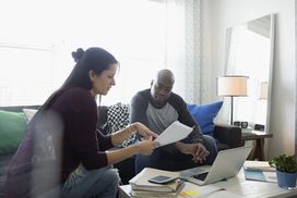 Couple sitting on a sofa with a coffee table holding files and a laptop balancing their checking account