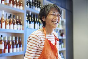 An employee of a wineshop stands in front of shelves of bottles and smiles.
