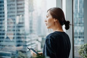 Young urban businesswoman using smartphone in the office in front of windows overlooking the city