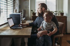 father sitting at desk trying to work with son on his lap crying