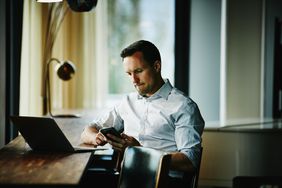 Businessman looking at smartphone while sitting at counter in office coworking space working on project on laptop