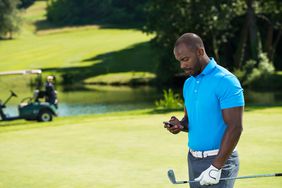 Golfer on sunny course holds club and looks at smartphone