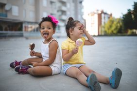 Two little girls sit on the sidewalk and enjoy melting ice cream novelties on a hot day. One of them appears to be experiencing "brainfreeze."