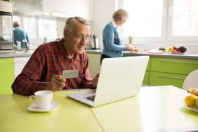 Smiling senior man on a laptop in home kitchen with credit card in hand
