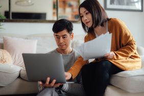 A couple sitting in a living room going over documents while sitting in front of an open laptop and looking intently at the screen