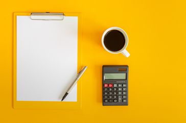 Top-down view of a clipboard with a blank white sheet of paper, a calculator, and a cup of coffee on a yellow background.
