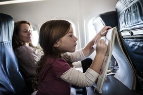 A child seated in an airplane passenger works on a coloring book while a woman seated beside her looks on.