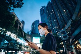 Young Asian woman with protective face mask using smartphone on the go, against illuminated neon commercial signs in city street in downtown district at night. The new normal of everyday life. Business on the go