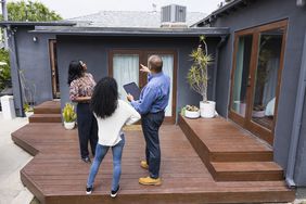A real estate agent shows the features of a home to two women. All are standing on a backyard deck and looking at the roofline, their backs facing the camera.