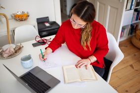person in red sweater writing notes from a book at a desk