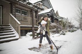 Woman wearing a baby in a baby carrier, shoveling snow outside a house