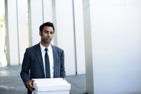 Young man in suit and tie walking out of a white building with a covered box after a layoff