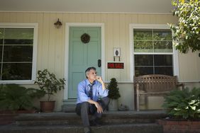 Man in suit sits on porch near for sale sign
