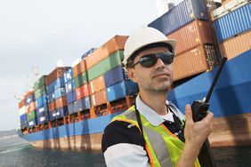 A worker in a hardhat holds a communication device and a cargo ship is in the background.