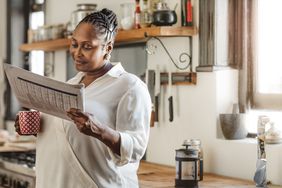 Woman reading a newspaper in her kitchen while holding a cup of coffee
