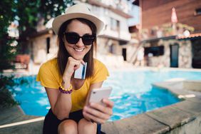 A woman sits in the shade near a sparkling swimming pool holding a credit card and using a smartphone