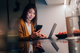 Young Asian woman looking at smartphone while working with laptop in the kitchen at home in the evening