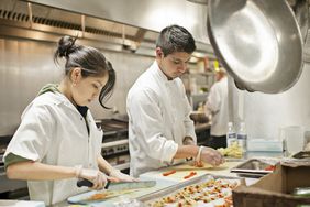 Young chefs in a gleaming professional kitchen prepare delicate-looking appetizers.
