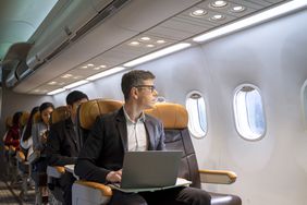 A man sits on a plane with a laptop on his lap looking out the window