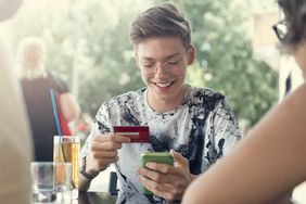 person with glasses wearing tie dye shirt, holding credit card and iPhone