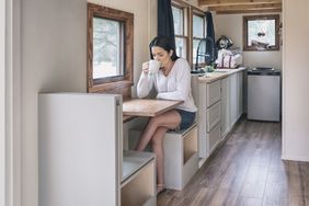 Woman sits at a small table drinking coffee in a tiny home. 