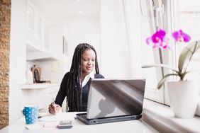 A woman with dreadlocks sits at a kitchen table, doing her income tax on a laptop 