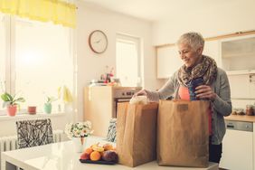 Woman stands smiling at paper shopping bags filled with groceries on kitchen table