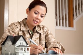 Woman in army fatigues signing check with miniature house in foreground