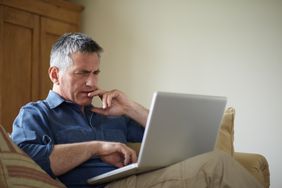 A man seated on a sofa looks quizzically at a laptop on his lap.