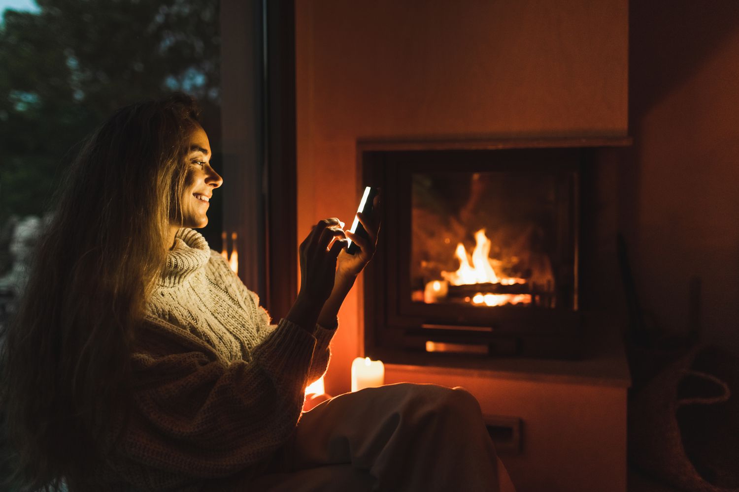 Smiling woman sitting with mobile phone on windowsill near fireplace at night