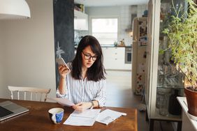A woman takes stock of her assets with phone, coffee, paperwork at home table
