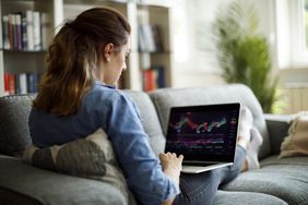 A woman sits on a couch reviewing trades on a laptop