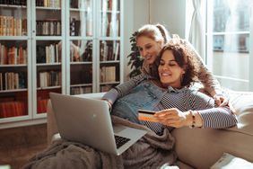 A female couple shops online while sitting on the couch with a credit card and laptop