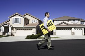Realtor walking in front of homes with 'For Sale' sign under his arm