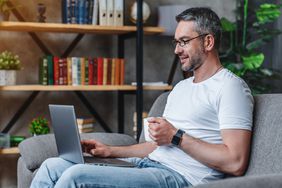 Man sitting on a couch with coffee mug in hand working on a laptop