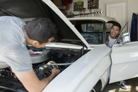 A son in the driver’s seat laughs at his father who is under the hood of the salvage car they are rebuilding