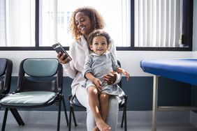 Mother using smartphone while toddler sitting on lap at doctor's office