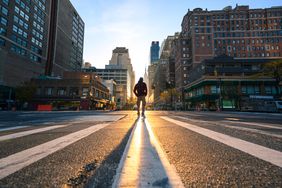 A person in a plaid jacket and fedora stands in the middle of a New York intersection, lit by the sun shining down the street behind them