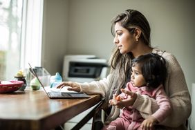Young mother multitasking at home on laptop with infant daughter